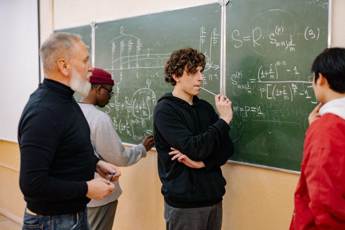 Students standing in front of a chaulk board. Two students are talking to the teacher and one is working a math problem on the board.